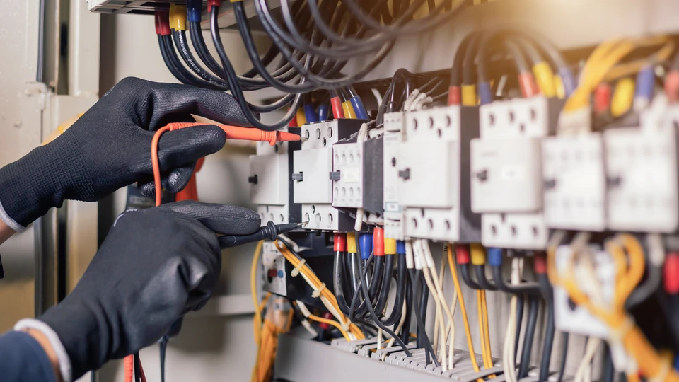 Un électricien en gants de sécurité noirs travaille sur un tableau électrique industriel en intérieur