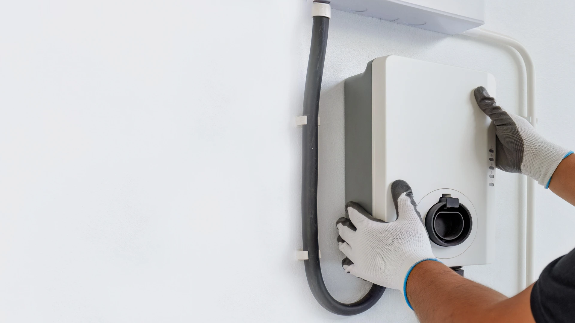 Électricien en tenue professionnelle installant une borne de recharge IRVE murale blanche ou grise sur la façade extérieure d'une maison résidentielle. La borne est clairement visible, fixée au mur en plein jour, avec des outils de travail visibles. L'électricien effectue les derniers branchements ou finitions.